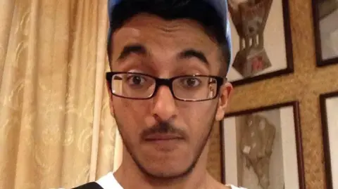 Mazin Kaladi looks at the camera. He has dark facial hair and is wearing a blue baseball cap and dark glasses.