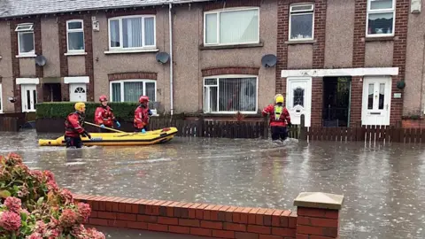 Emergency services staff take an inflatable boat to rescue people. The street is flooded with water which goes above the knees of rescue workers.