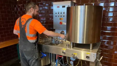 Alex Pope/BBC A man, in overalls, holding on to metal machinery inside a brewery. He has his hair tied back and his back to the camera. There are red glazed tiles behind him. 