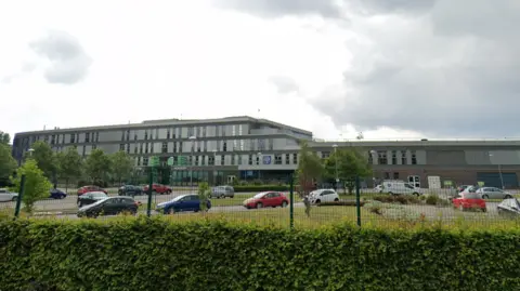 Google A Google streetview image of a school building which is set behind a green mesh fence and large green hedge. A number of cars are parked in front of the large grey building which features rectangular windows across three floors.