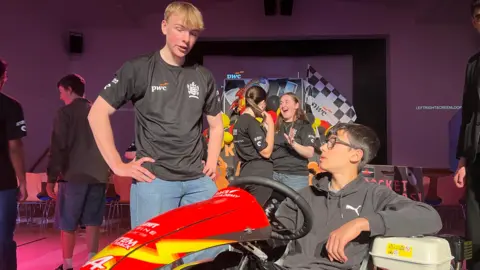 Two people interact beside a red go‑kart; one is seated in the kart while the other stands nearby wearing a black PwC T‑shirt, with event staff and displays on a stage in the background