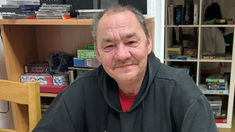 A man in a room, wearing a black top with a red top underneath. He has a plate of food in front of him that is half eaten, He is sitting at a table, with a chair next to him and items behind him. He is smiling and looking straight at the camera. 