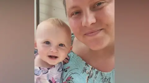 Handout A mum poses for a selfie with her baby daughter. The mum has brown hair which is tied back and is wearing a light blue top with a floral pattern on. Her baby is smiling, has very short blonde hair and is wearing a pink, purple and white dress with butterflies.