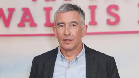 Steve Coogan stands in front of a white wall. He is wearing a blue dress shirt and a grey jacket. 