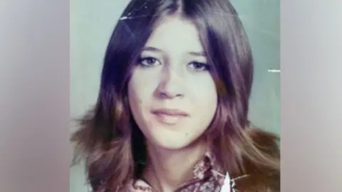 Laura Aimee, in a school photo from the 1970s. She has long brown hair parted down the middle and brown eyes, and wears a patterned blouse. 