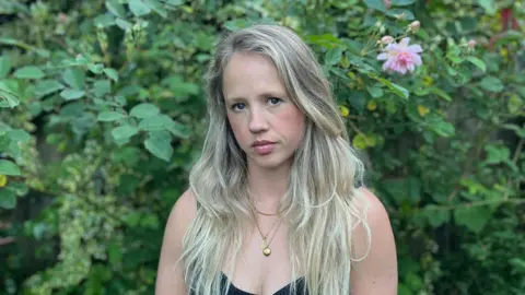 Kiri Nowak Kiri Nowak staring into the camera, with a rose bush in the backgroundbush in the backg