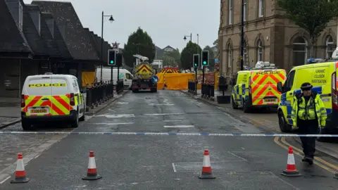 A street which has been cordoned off with police tape, three police vans are inside the cordon and a police officer stands guard. There is a road sweeper in the background and several buildings.