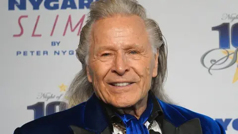 Peter Nygard smiles at the camera at a promotional event. He has long white-grey and wears a blue velvelt suit jacket. 