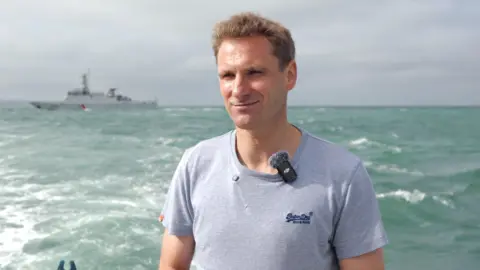Jack Valpy/BBC MP Chris Philp, in a grey t-shirt, stands on a boat in the sea. A large, grey ship is visible in the background.