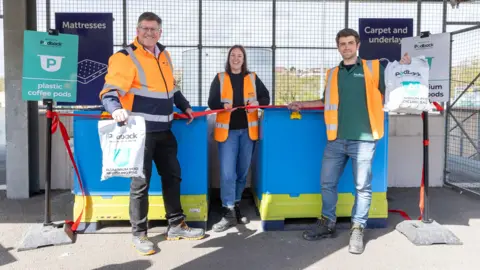 Three people wearing orange high-vis jackets standing in front of a red ribbon holding bags that say "Podack" on them. They are also standing in front of two blue and yellow coffee pod waste bins.