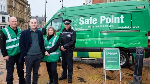 Wakefield Council Two people in green vests, a man in a black suit and a police officer in uniform stand in front of an ambulance, which has been painted green, and says "safe point" on the site in large white lettering.