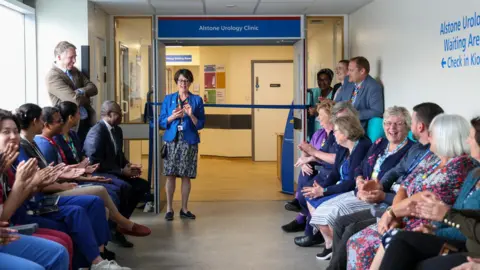 Gloucestershire Hospitals Trust Grand opening of new urology clinic at Cheltenham General Hospital, with people clapping as a woman prepares to cut the ribbon