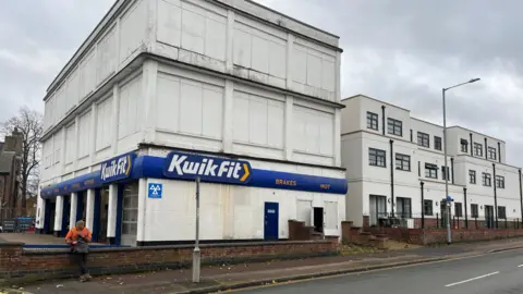 Clare Worden/BBC A three-storey tall white building with the windows covered with white. There is a large blue Kwik Fit sign on the ground floor. 