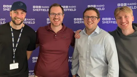 Steve Jones/BBC Four men stood in front of a purple BBC Radio Sheffield banner. They are all over 6ft in height and smiling in the pitcure. Two of them have dark hair and glasses, another wears a baseball cap while the fourth man has medium-length blonde hair.