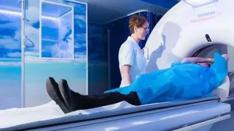 Person dressed in a blue gown lying down with arms raised about to enter a hospital scanner. A nurse is standing next to the machine dressed in white.