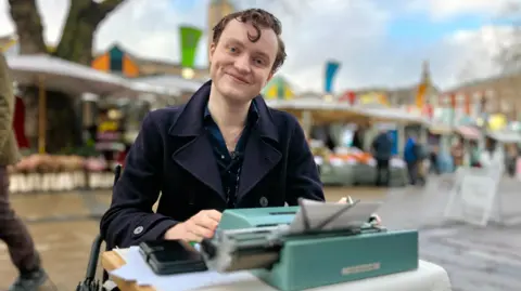 Nathan Rodney-Jones is in a blue or green shirt, and wearing a navy coat over the top. He has a typewriter on a table. He has curly brown hair. There are market stalls behind him. There are some blue skies above, but also white cloud.