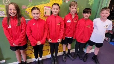 BBC Six pupils stand side by side in front of a green wall. Five of them are wearing bright red jackets while the sixth is in a white T-shirt. 