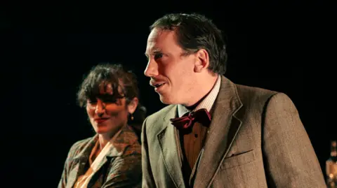 A man and woman, in 1950s style clothing, are looking to the left of the frame and standing in front of a black background.