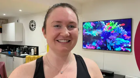 BBC/Andy Watson Lauren Emmerson wears a black exercise top with her hair in a pony tail. She is standing in front a a tv which has a fish tank screen saver. 