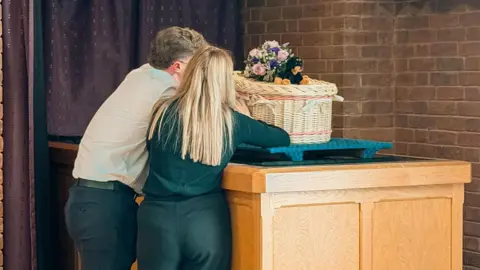 Jade Morris Two people with their backs to the camera, stand next to a baby's wicker coffin, which is on a waist-high plinth. There is a bouquet of flowers on top of the coffin.