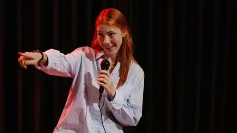 A woman with long ginger hair holds a microphone in one hand and points forward with the other. She is wearing a blue and white striped shirt and is stood in front of a black background.