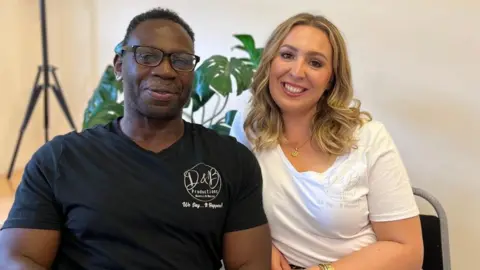 A man and woman are smiling and sitting next to each other with plants behind them.