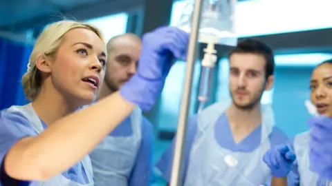 A nurse with tied-back blonde hair and blue scrubs holding the top of an intravenous drip. Three other nurses dressed in scrubs - two men and a woman - stand behind watching.