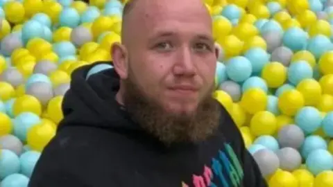 Chad Allford in front of a ball pit, wearing a black hoodie. He has short hair and a brown beard