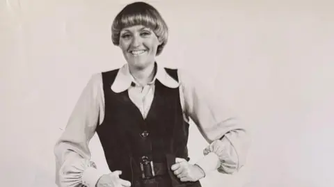 Supplied A black-and-white photo of a younger Mildred wearing a blouse with large collars and a velvet waistcoat. She has her hands on her hips and is smiling at the camera