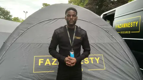 A man standing infront of a van and infront of a tent. He is wearing all black and the van behind him is matt black. There are yellow bright signs that say in capital letters FAZAMNESTY