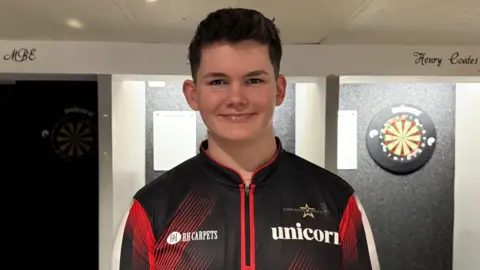 A young man with dark hair stood in front of a darts board.