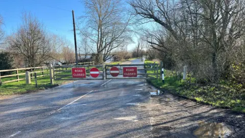 John Devine/BBC Rural scene with some metal barriers closed across a road with no entry signs and the words "road closed" in white on a red background, a small amount of water appears to be covering the road. Trees and telegraph posts can be seen too.