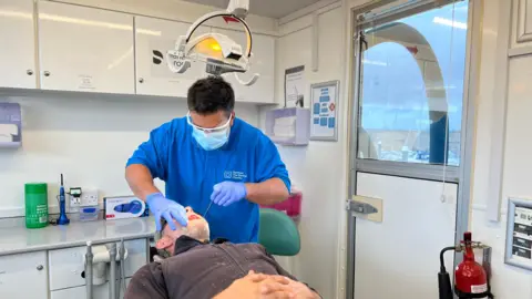 A dentist looks into a fisherman's mouth. The fisherman is lying down on dentist chair which has been reclined and has his mouth open. He is wearing a grey zip-up fleece and has grey stubble on his face. The dentist is wearing a blue short sleeved top with medical gloves, a face mask and special dental glasses. He is using dentists' tools to see into the fisherman's mouth and an overhead lamp is shining down on them.