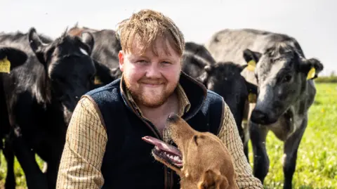 Kaleb looks into the camera smiling, with a dog looking up at him and four cows behind him