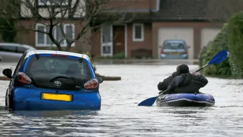 A resident uses a boat to make their way through floodwater in Chertsey in 2014. Residential houses can be seen in the background, while cars are also surrounded by water.