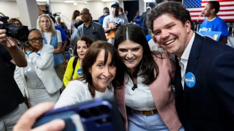 Getty Images Three people are taking a selfie in a crowded room. On the left of the three is a middle aged woman holding the camera. In the middle is Haley Stevens with a broad smile. On the right is Gary Peters dressed in a dark suit and open necked, white shirt.