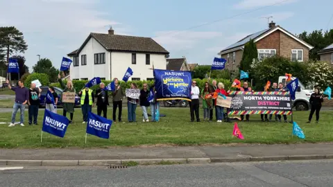 BBC/Seb Cheer A group of 18 people stand with flags on a patch of grass in front of some houses, holding flags and banners reading "NASUWT Yorkshire and the Humber" and "National Education Union North Yorkshire District".