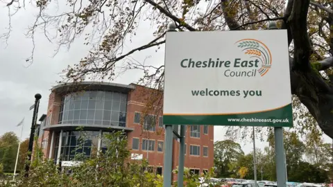 A sign for Cheshire East council outside of a council building. A tree can be seen hanging over the sign. 