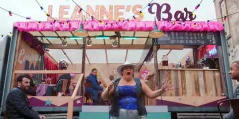 BBC/Expectation TV Leanne (Jayde Adams) is singing, wearing a bluish  corset benignant   top, with a gilet, metallic  jeans and a achromatic  cowgirl hat