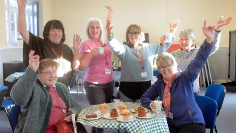 Positive Community Action Six adults, two sitting and four standing wave their arms in the air for a posed photograph taken in a community cafe. On the table in the centre of the photo are a number of cakes and muffins. A window can be seen to the far left.