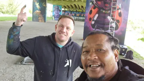 Tony Nero Nathan and Tony, both men, smiling for camera while taking a selfie in front of the artwork.