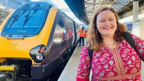 Woman - Aimee Smith - in bright maroon dress, smiling at camera, on rail platform, with train in the background.