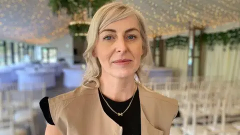 A blonde woman looking at the camera, she is wearing a tan brown sleeveless coat and black top