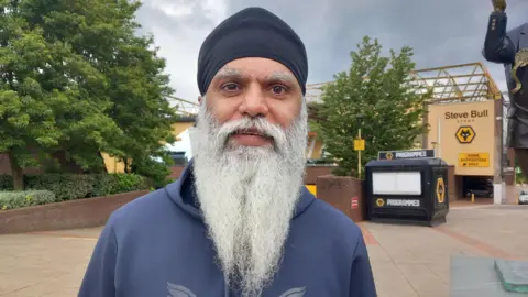 Manny Singh Kang Manny Singh Kang smiling in front of Molineux Stadium wearing a blue hoodie 