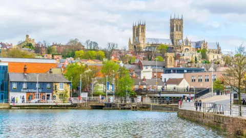 Brayford Pool with Lincoln cathedral on the hill in the background. The Royal William IV pub is on the riverside on the left and a road runs along the bank to the right.