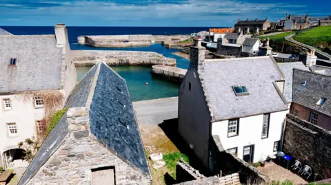 Getty Images A view across Portsoy's Old Harbour in Aberdeenshire. There are rows of traditional buildings with peaked, slate roofs. The harbour has low, stone walls. It is a bright sunny day.