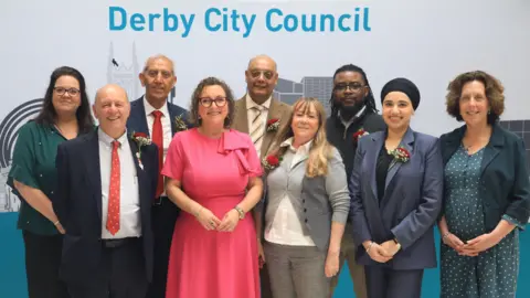 A group of councillors stood smiling infront of a board saying 'Derby City Council'
