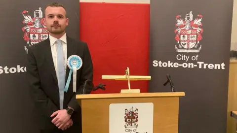 A man stands next to a podium at a by-election count. He has his hands clasped in front of him and he's wearing a blue and white Reform rosette. 