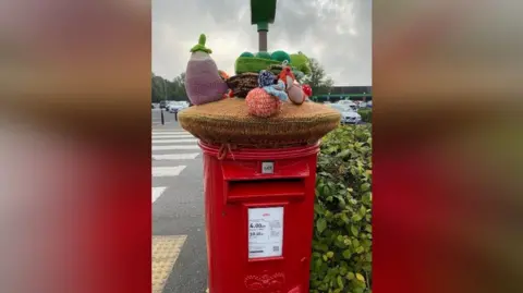 Whinfield Yarn Bombers A photo of a red post box with a knitted topper featuring different fruit and veg.