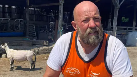 Paul Costain Paul, who has a bald head and a beard, stands in his farm with a sheep behind him - he is wearing a white T-shirt and orange vest